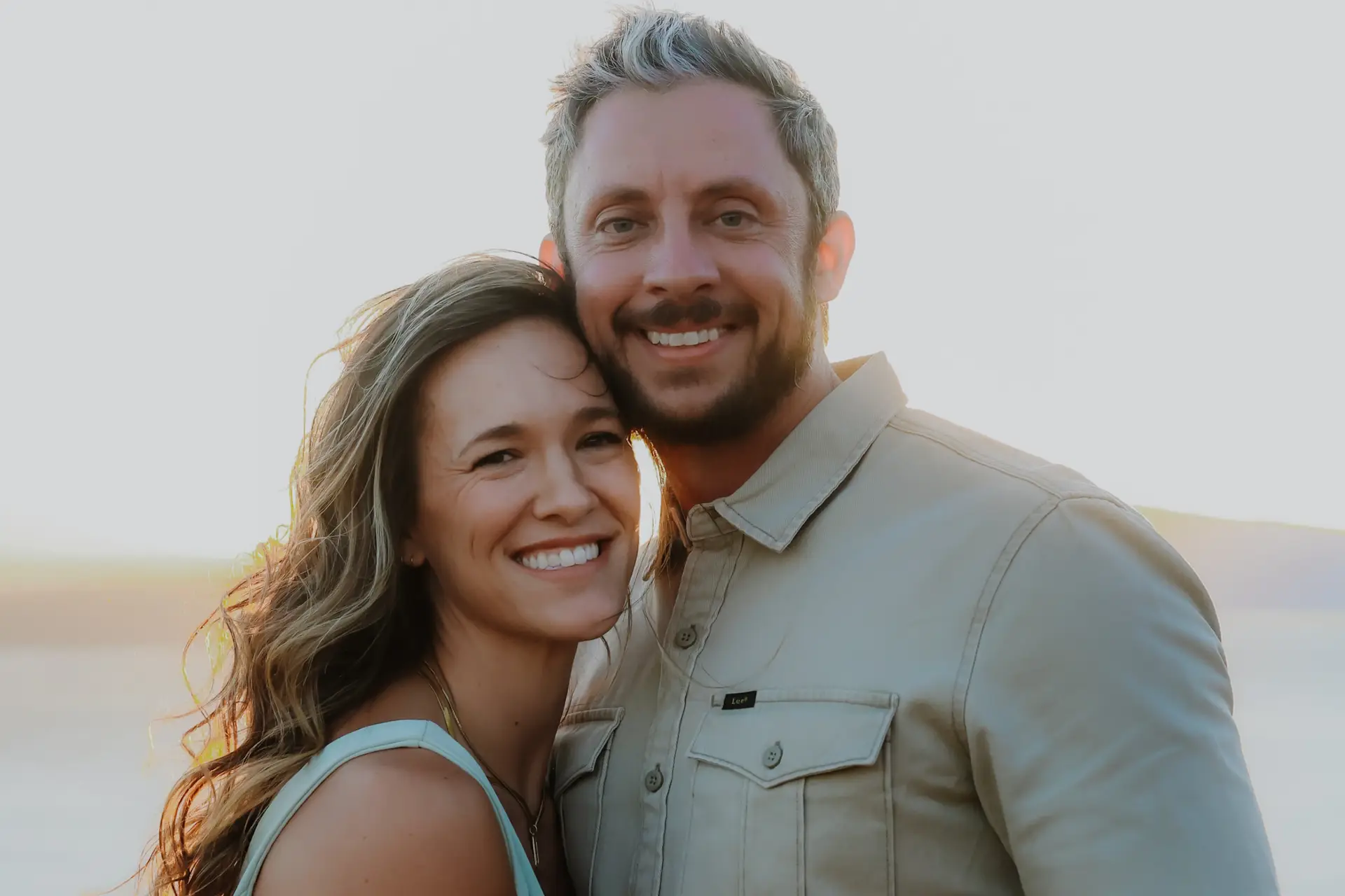A smiling couple embraces outdoors at sunset, faces close together with warm light on their skin.
