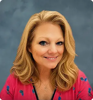 Head-and-shoulders portrait of a smiling blonde woman in a pink cardigan against a blue backdrop.