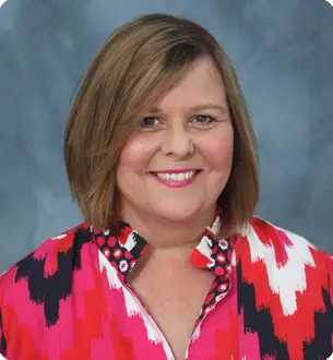 Smiling woman with shoulder-length light brown hair wearing a colorful red-pink patterned blouse against a blue backdrop.