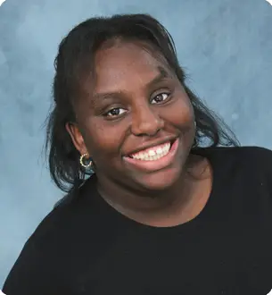 Smiling woman with dark hair and gold hoop earrings wearing a black top against a blue studio backdrop.