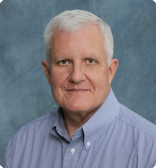 Portrait of a smiling older man with white hair in a light blue shirt, against a blue background.