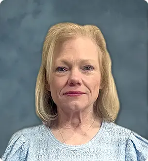 Head-and-shoulders portrait of an older woman with blonde hair, wearing a light blue blouse, neutral expression, blue-grey background