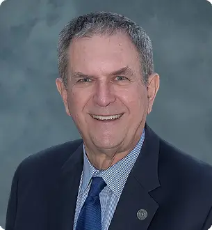 Head-and-shoulders portrait of a smiling older man in a navy blazer, light blue shirt, and blue tie against a blue-gray backdrop.