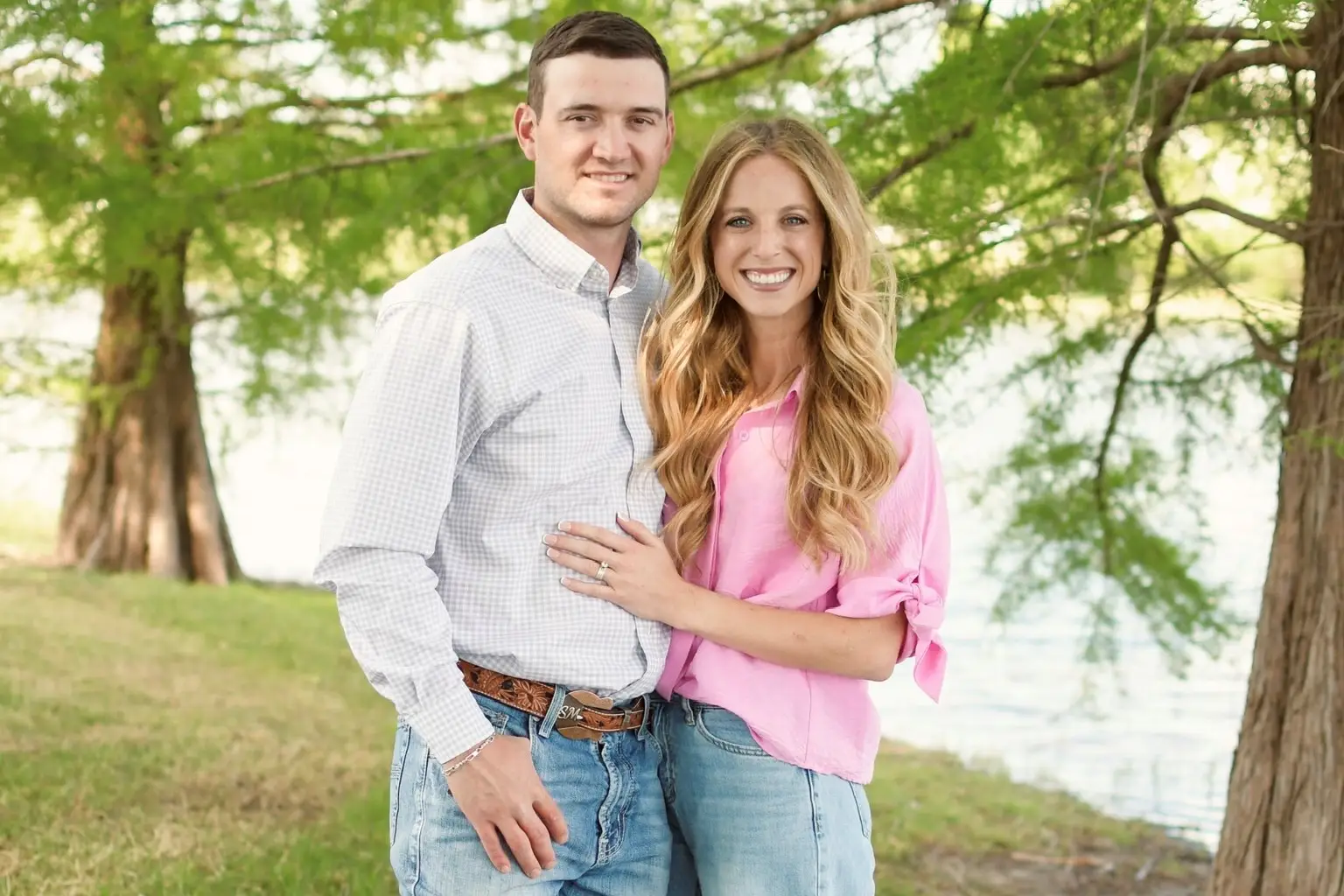 Smiling couple standing together outdoors by a lake with trees in the background.