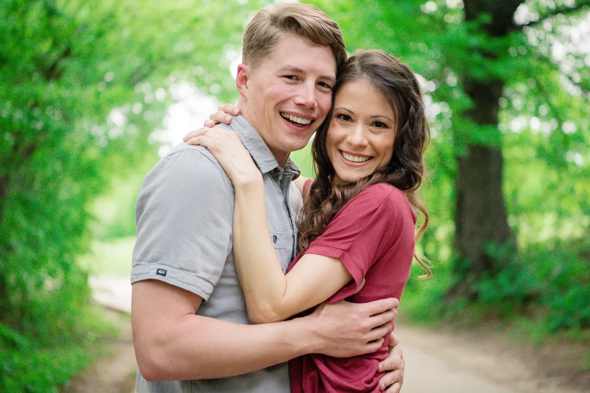 Happy couple hugs in a sunlit park, smiling at the camera with green trees in the background.