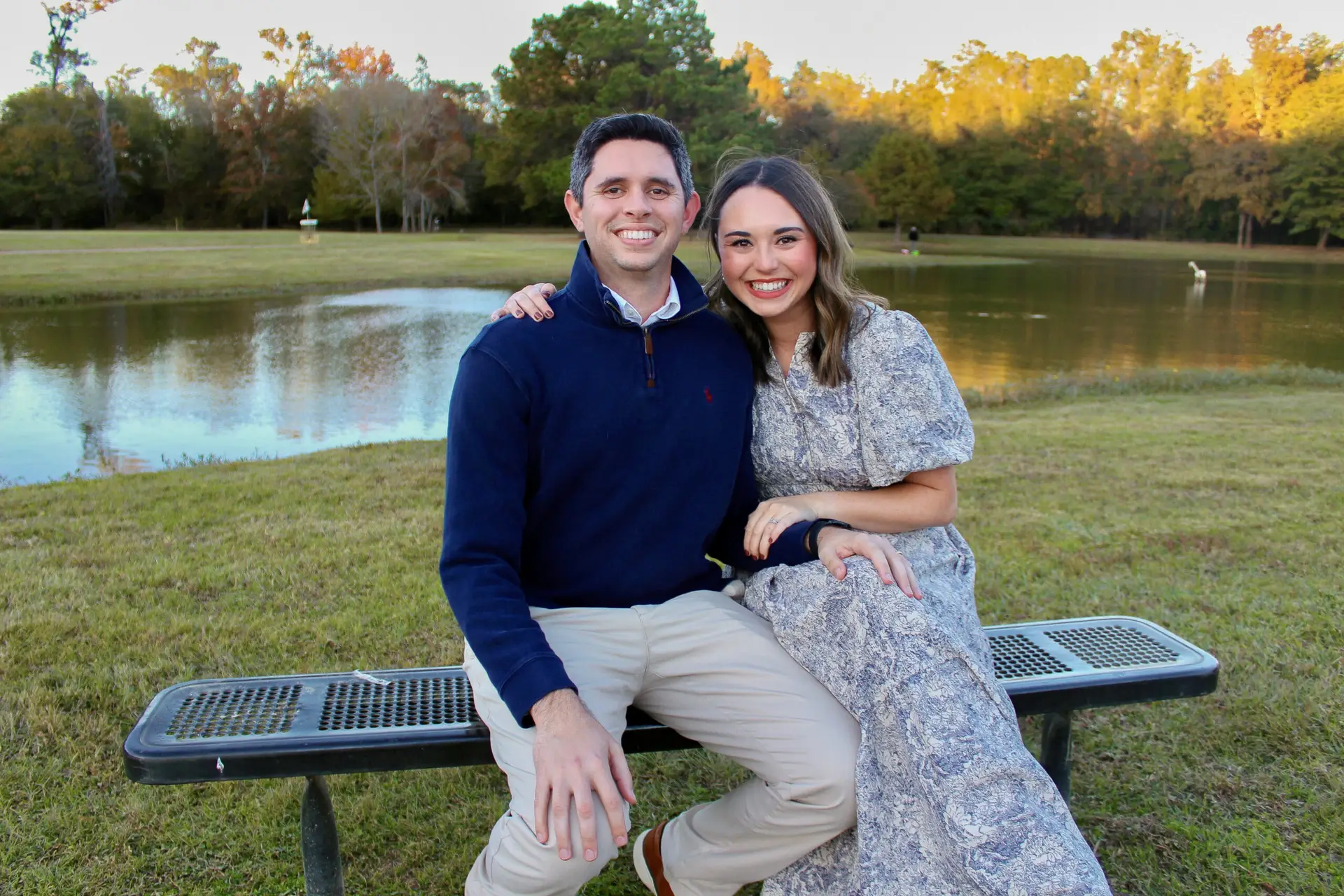 Smiling couple sitting on a park bench by a pond with autumn trees in the background.