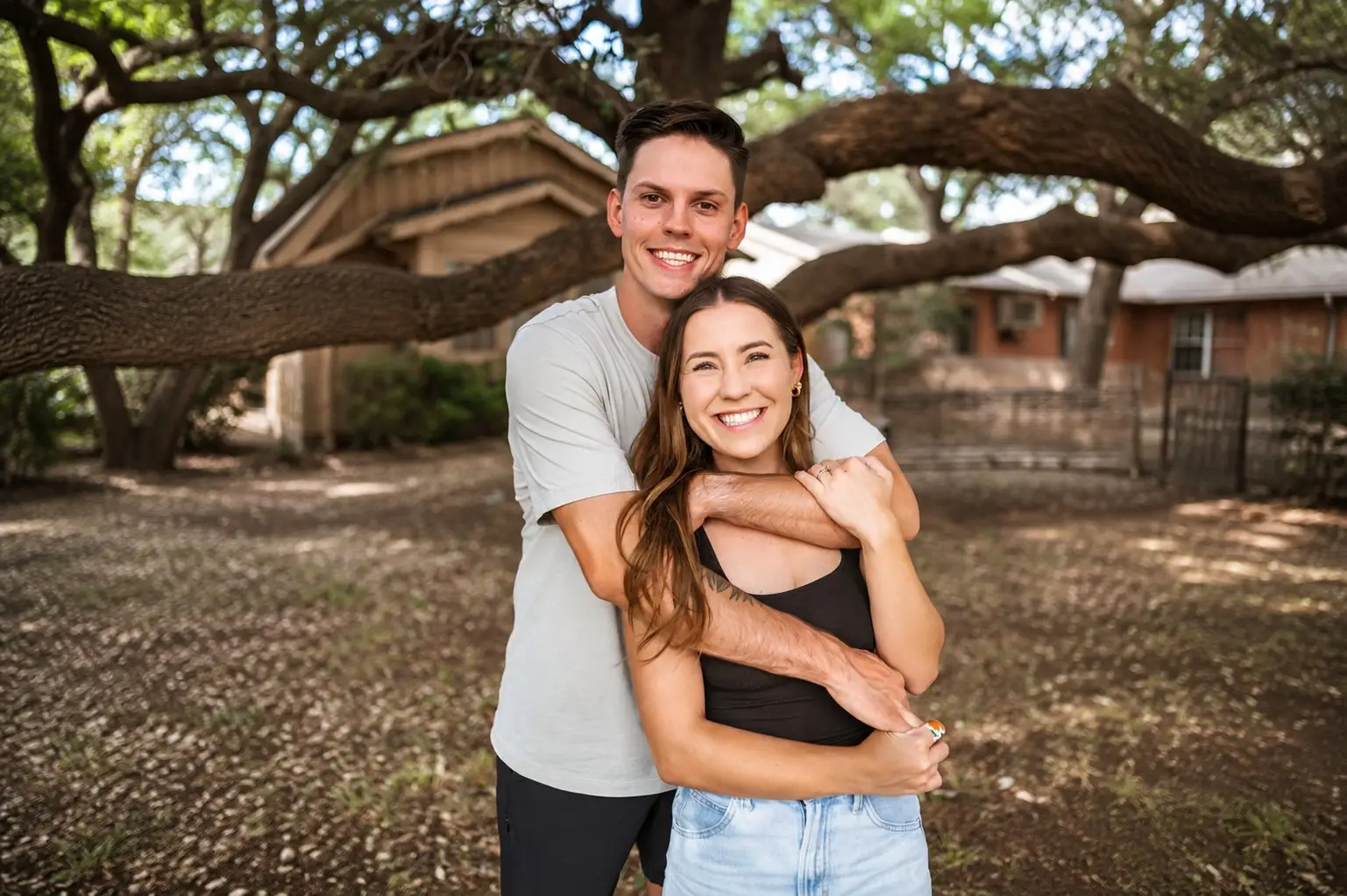 Smiling couple embracing outdoors under a large tree in a residential yard.