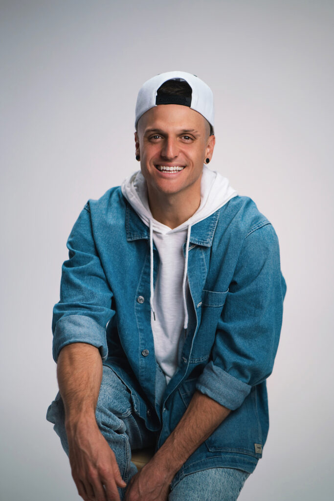 Young man in a denim jacket and white hoodie, wearing a backward white cap, smiling at the camera.