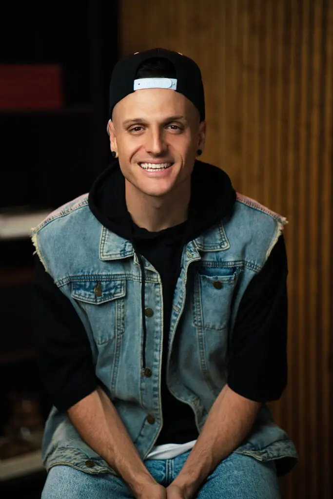 Smiling man wearing a backward baseball cap, denim vest, and black hoodie, seated indoors against a wooden wall.