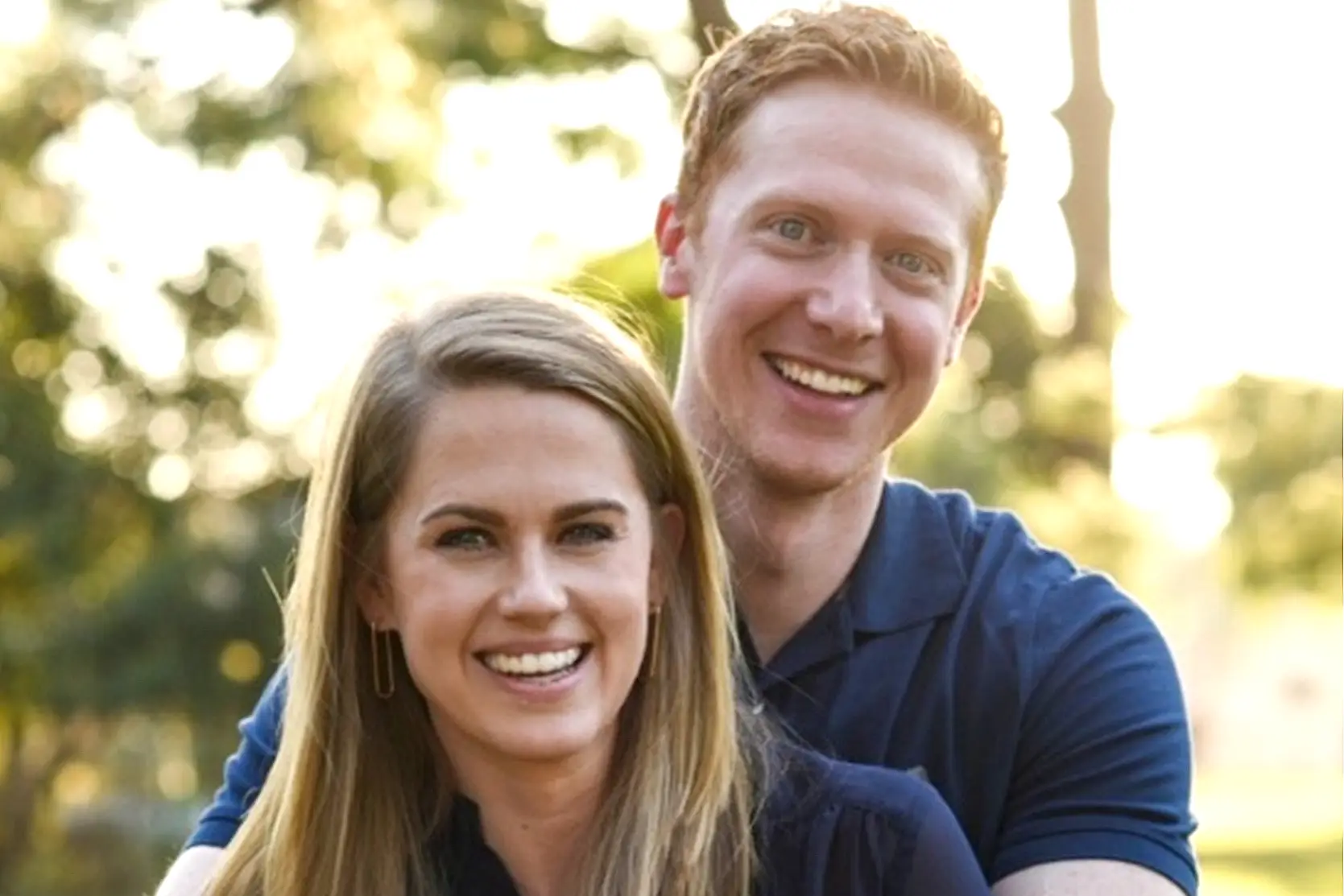 Smiling couple outdoors in warm sunlight, the man standing behind the woman with his arms around her shoulders in a park setting.