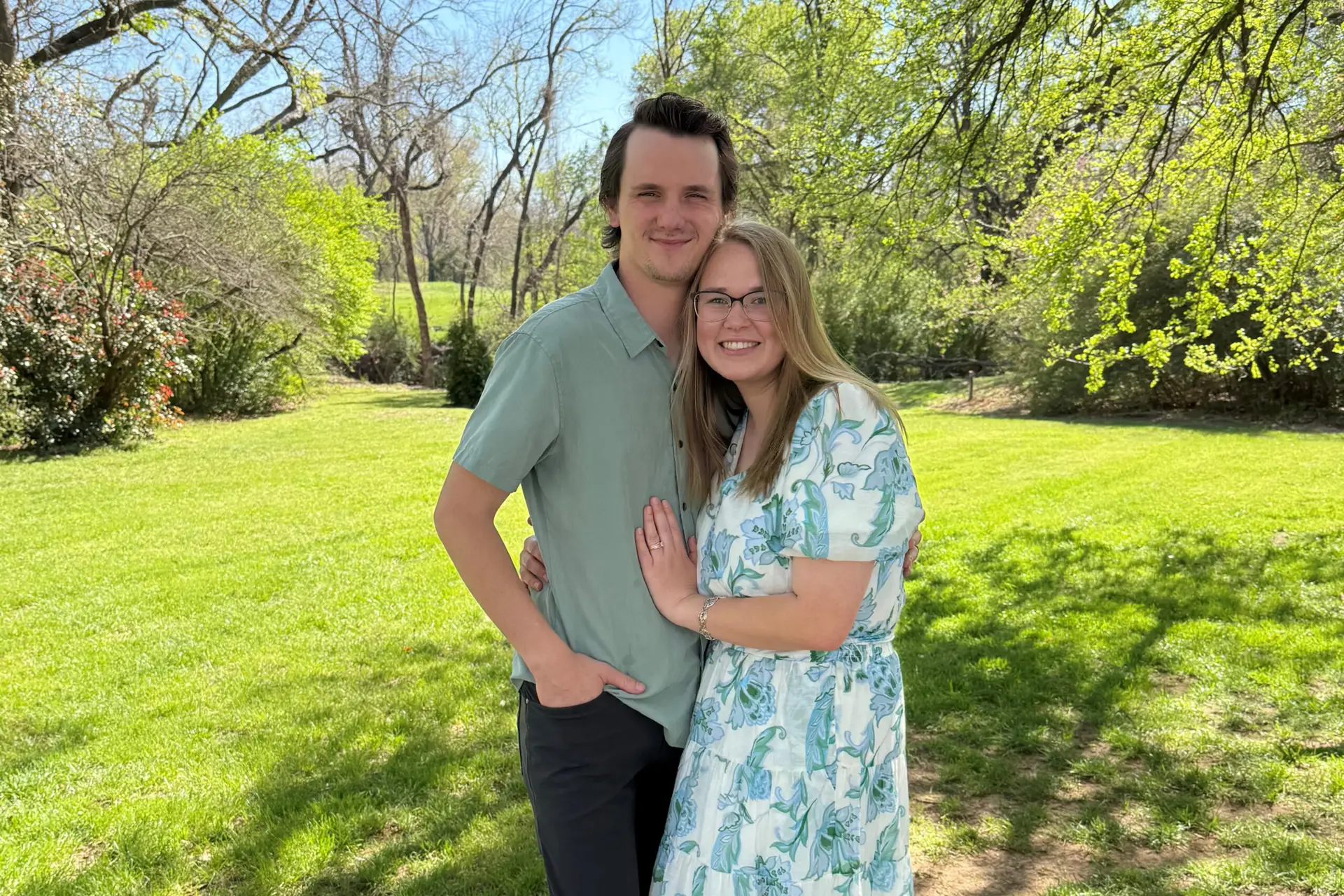 Smiling couple embracing in a sunny park with green grass and leafy trees in the background.