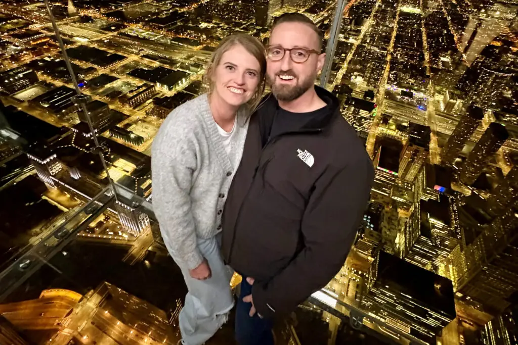 Couple posing on a glass observation deck above a city at night, smiling at the camera.