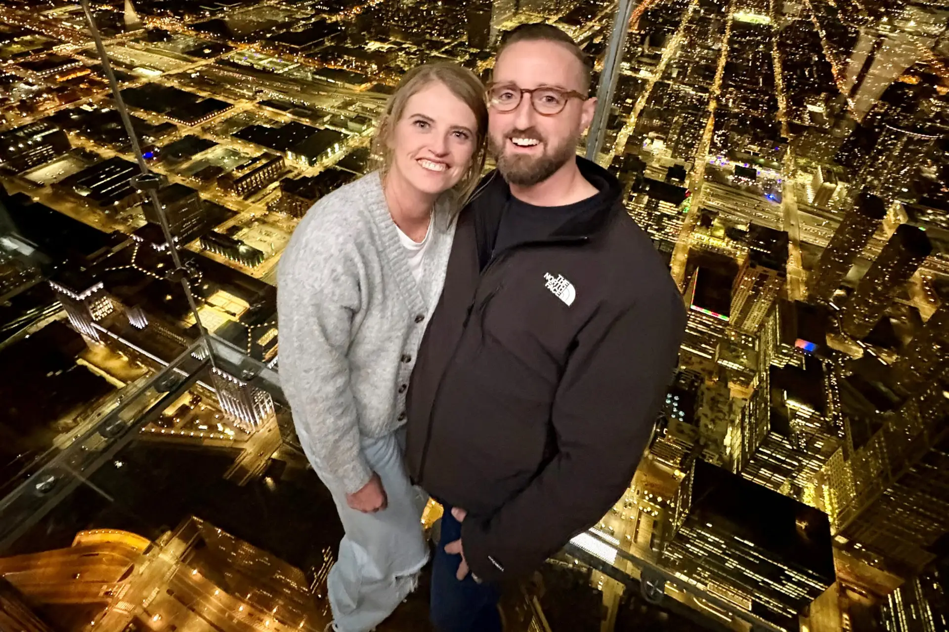 Couple posing on a glass observation deck above a city at night, smiling at the camera.