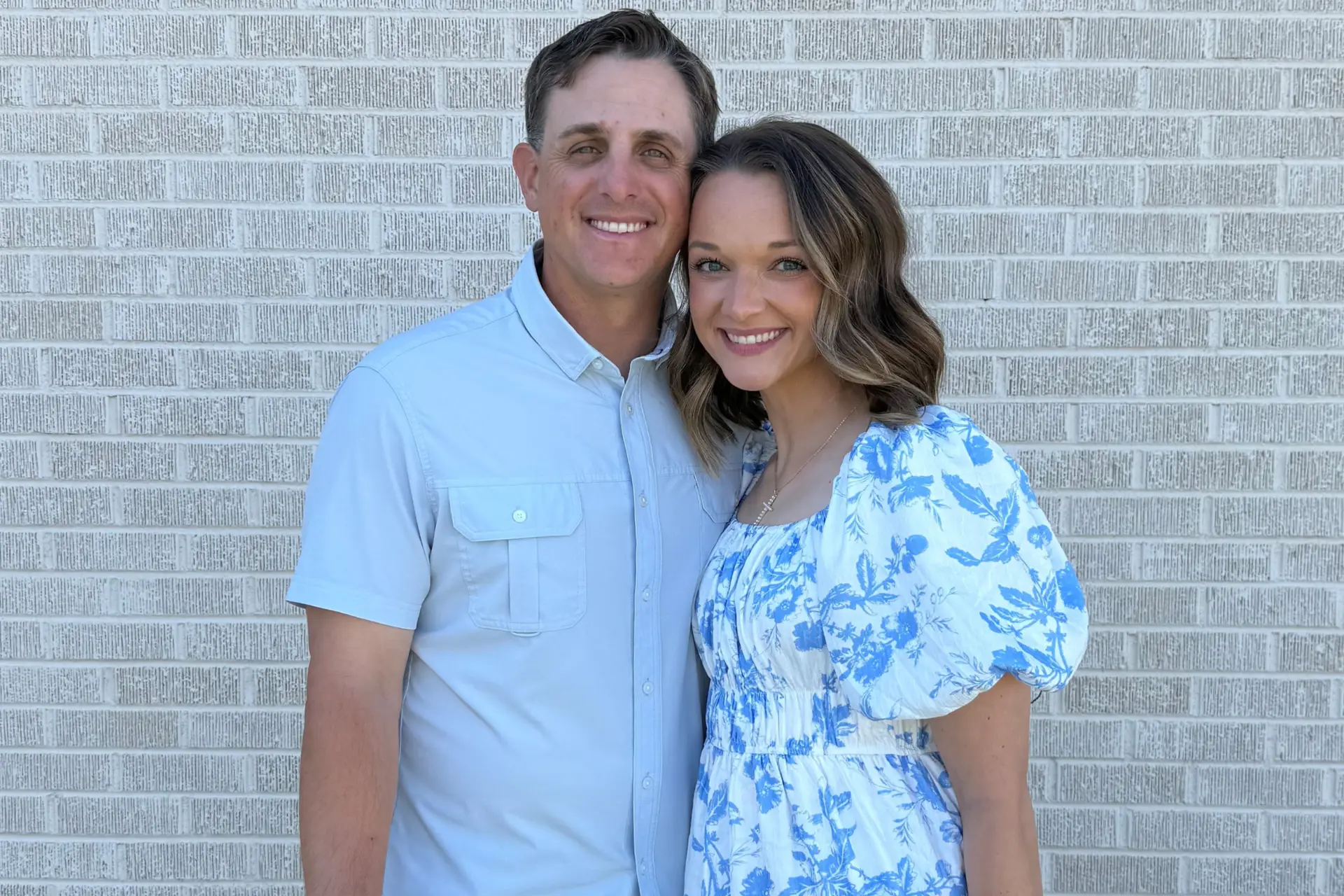 Smiling couple stands side by side against a light gray brick wall in casual blue outfits.